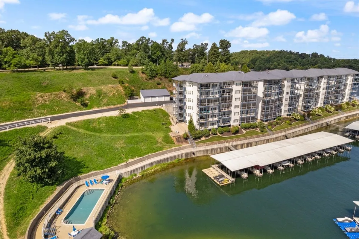 This image features an inviting swimming pool set along the waterfront, with a well-maintained apartment building and dock visible in the background. The pool area is equipped with lounge chairs, and the serene lake and dock highlight easy access to water activities and relaxation by the shore.