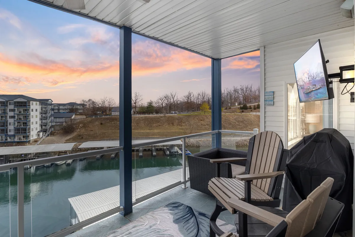 A tranquil balcony setting overlooking the water at sunset, with comfortable seating and a mounted television. The sky glows with sunset hues, inviting relaxation after a day spent outdoors.