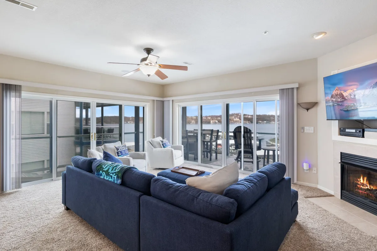 A spacious living room featuring a large blue sectional sofa positioned to enjoy both the warmth of a fireplace and spectacular floor-to-ceiling windows that frame serene lake views. The room also includes a television displaying mountain scenery, creating a cozy and inviting hub for relaxation and gatherings.