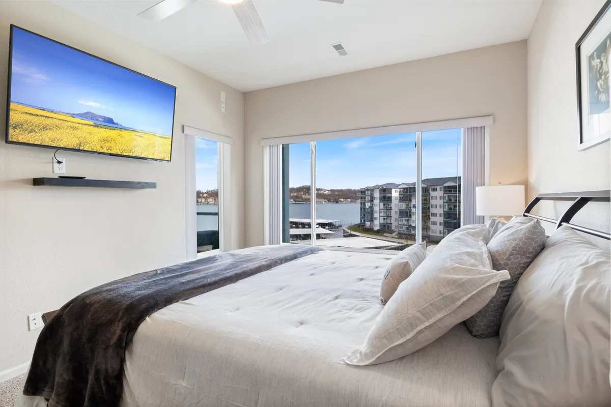 A peaceful bedroom with a large bed adorned with decorative pillows, a sizeable window that lets in natural light and offers a view outside, and a mounted TV displaying a scenic image. The tranquil decor and soft bedding set a restful, inviting mood.