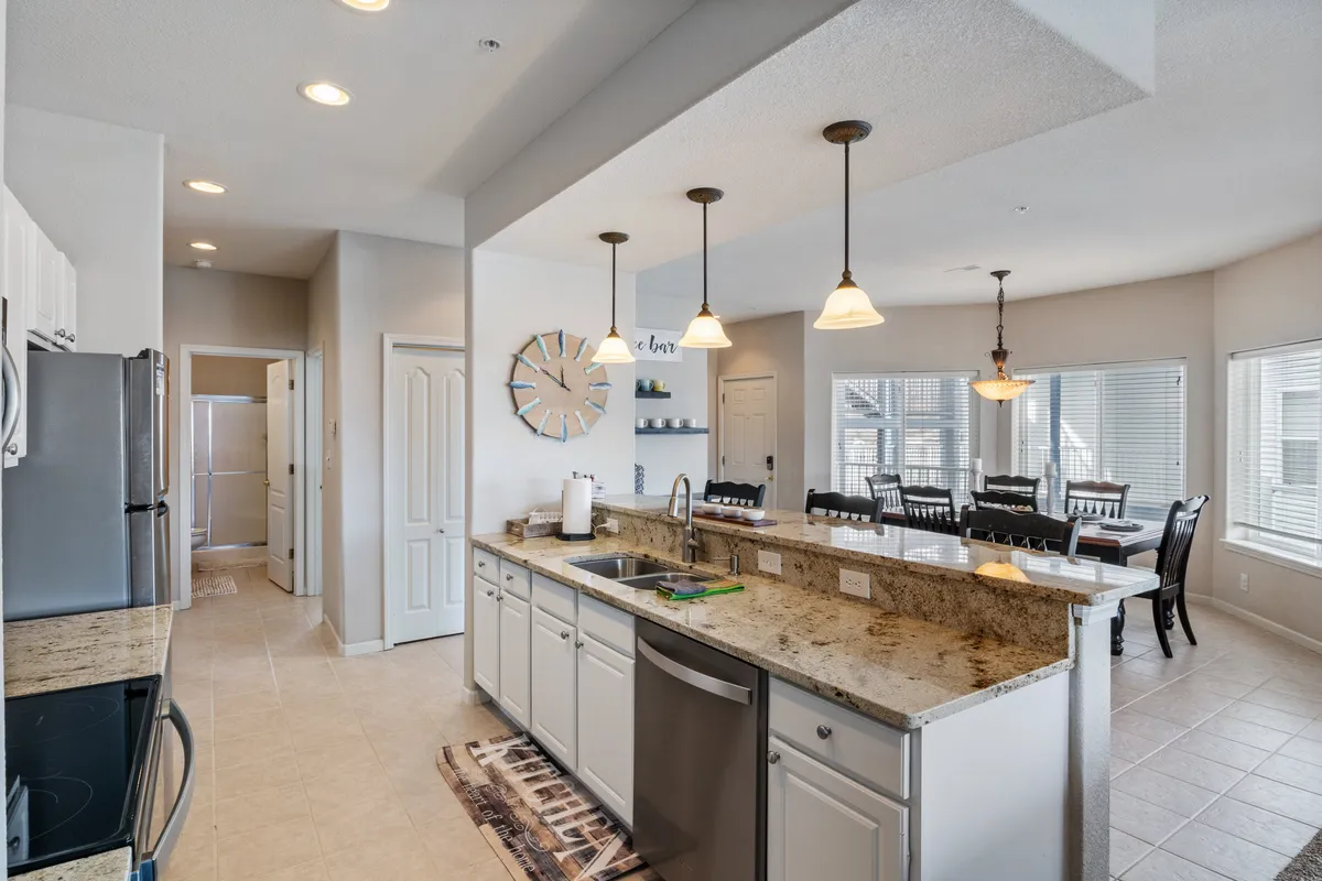A bright, inviting interior scene with a kitchen island connected to a stylish dining area. Large windows let in abundant natural light and offer glimpses of the lake beyond. The space is decorated with a decorative wall clock and is set up for socializing and entertaining.