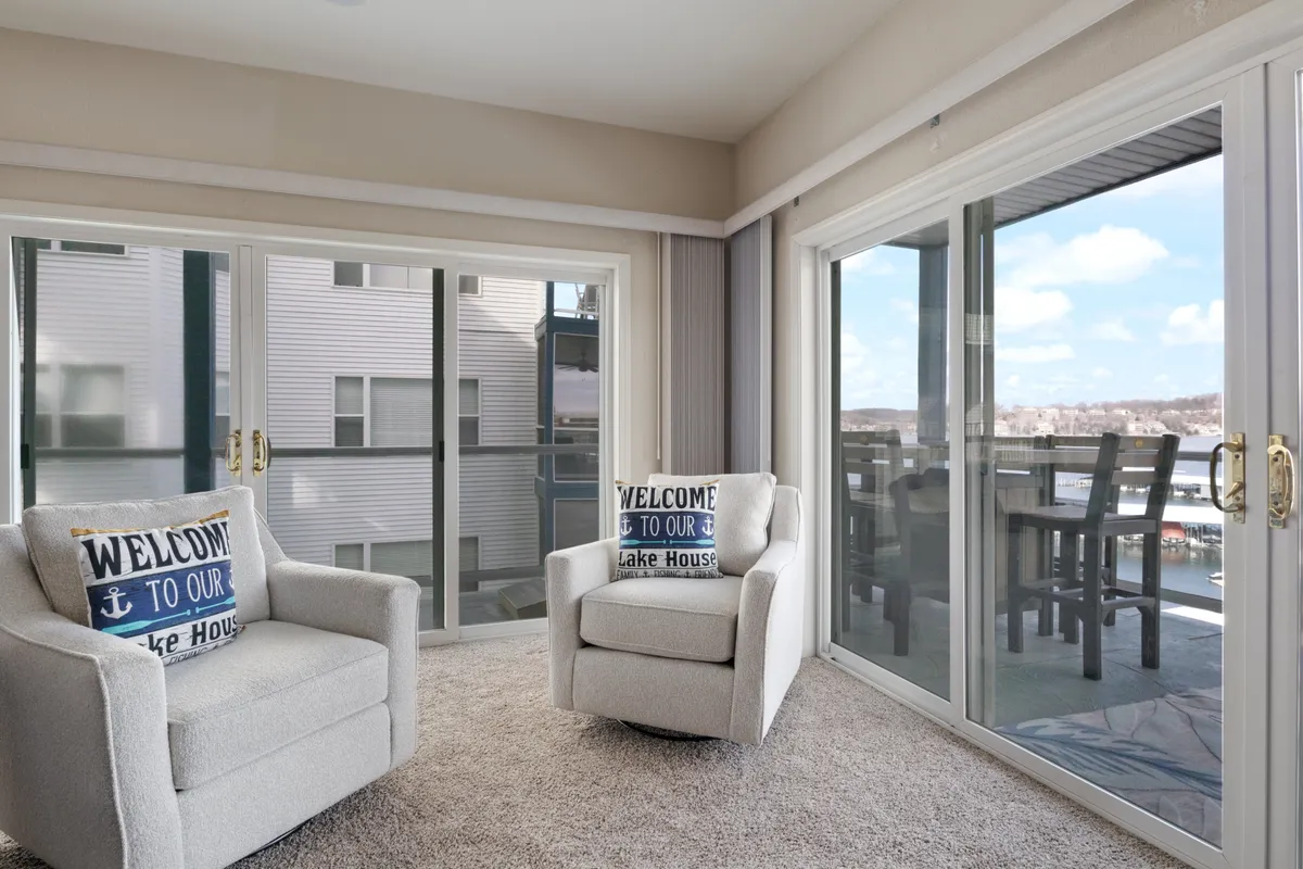A cozy seating nook with plush armchairs and decorative pillows featuring a welcoming message. Large glass doors open directly to the outdoors, creating a harmonious connection between indoor relaxation and the scenic lake setting.
