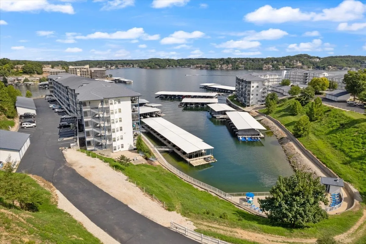 The image centers on the waterfront docks edged by lush landscaping and set against a clear blue sky, with a multi-story apartment building in the background. The docks are ready for boating, fishing, and lakeside picnics, supporting a variety of water-based activities.