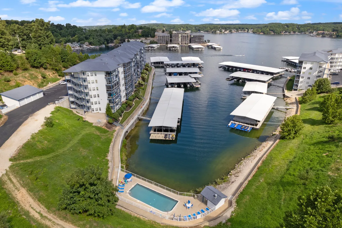 This vibrant exterior shot captures a waterfront scene featuring a marina and a swimming pool, with buildings lining the water's edge. The pool area offers lounge seating while the marina showcases boats docked by the shore, all set against the backdrop of the lake and a glowing sunset.