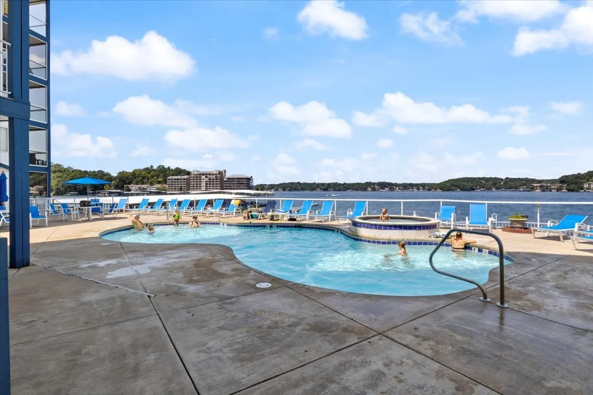 An inviting pool area with lounge chairs and umbrellas overlooks the serene lake, creating an ideal setting for sunbathing and socializing. The scene exudes a cheerful, leisurely vibe, with the water sparkling in the sunlight and open views drawing the eye to the horizon.