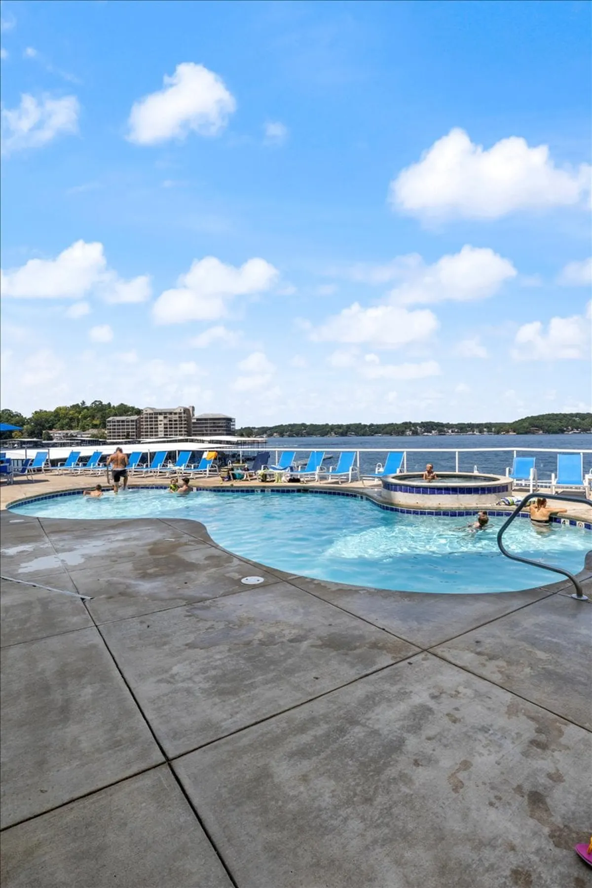 This lively scene captures a sparkling swimming pool overlooking the lake, with people enjoying the water and lounging poolside. The view extends over the water, creating a seamless transition from pool to lake and emphasizing the social, recreation-focused atmosphere.